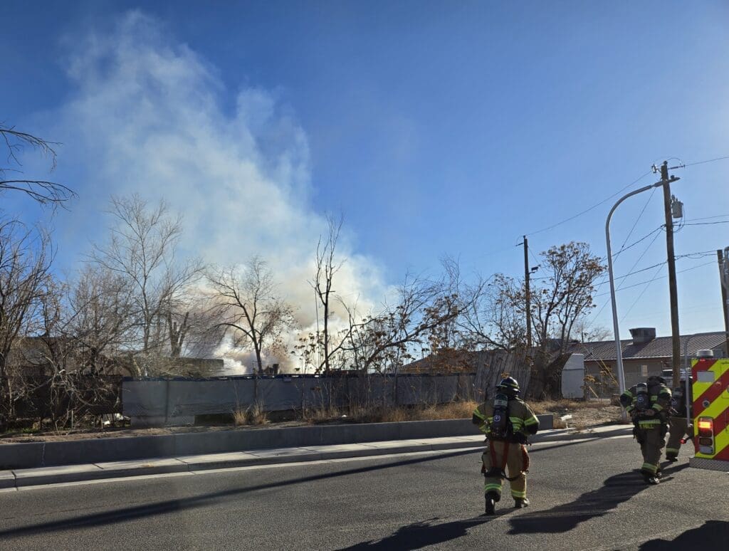 Three firefighters standing in street in front of a fence that had smoke coming out behind it.