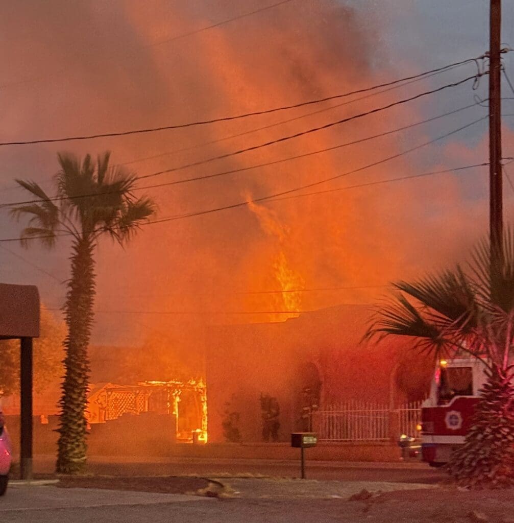 Palm tree in front of a house that is engulfed in flames, and a fire truck parked off to the side.