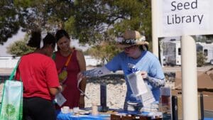 people talking in the shade with a sign in the photo that says seed library.