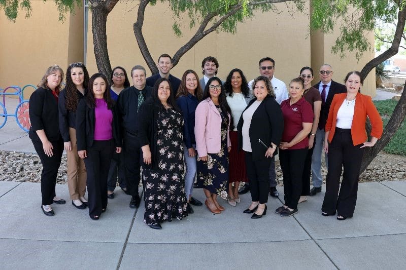 Group of people standing together on a sidewalk in front of a tree.
