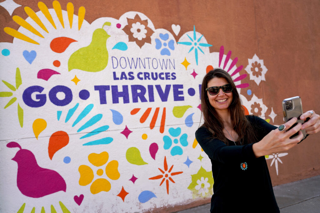 Woman with sunglasses taking a selfie in front of a downtown Las Cruces Mural