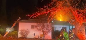 Firefighters standing in front of a house with flames coming out of the back of the house.
