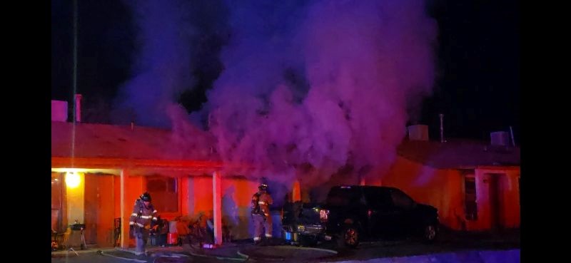 Firefighters standing outside a burning structure with a truck parked in front,