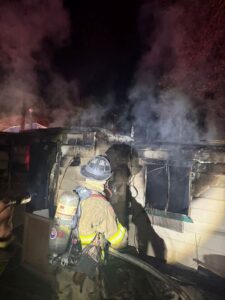 Firefighter in full gear working on a house with black smoke and busted out windows.