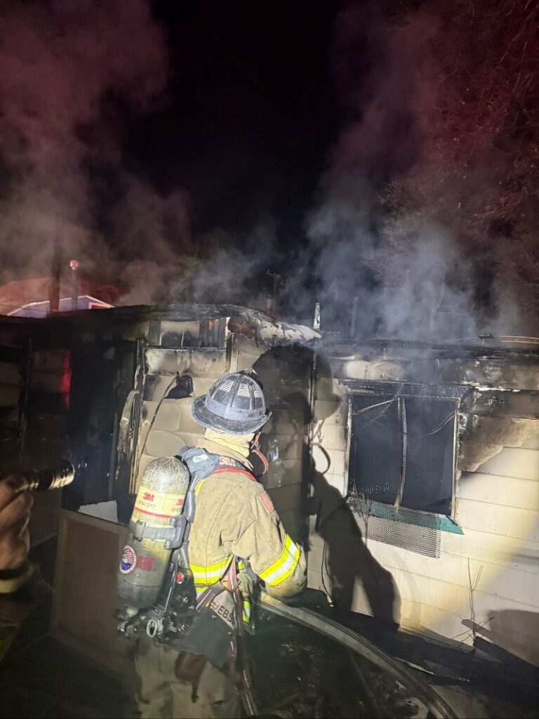 Firefighter in full gear working on a house with black smoke and busted out windows.