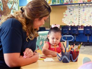 Woman with arms propped on a round table overlooking a young child coloring with colored pencils with a couple of cups on the table holing more pencils.