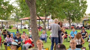 Photo of a couple dancing by a tree in the park with several other people sitting in folding chairs in the park on the grass.