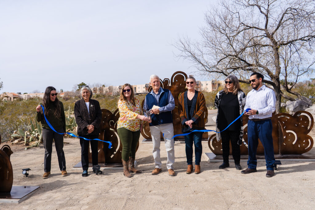 A group of people cut a blue ribbon in front of art sculptures at a park.