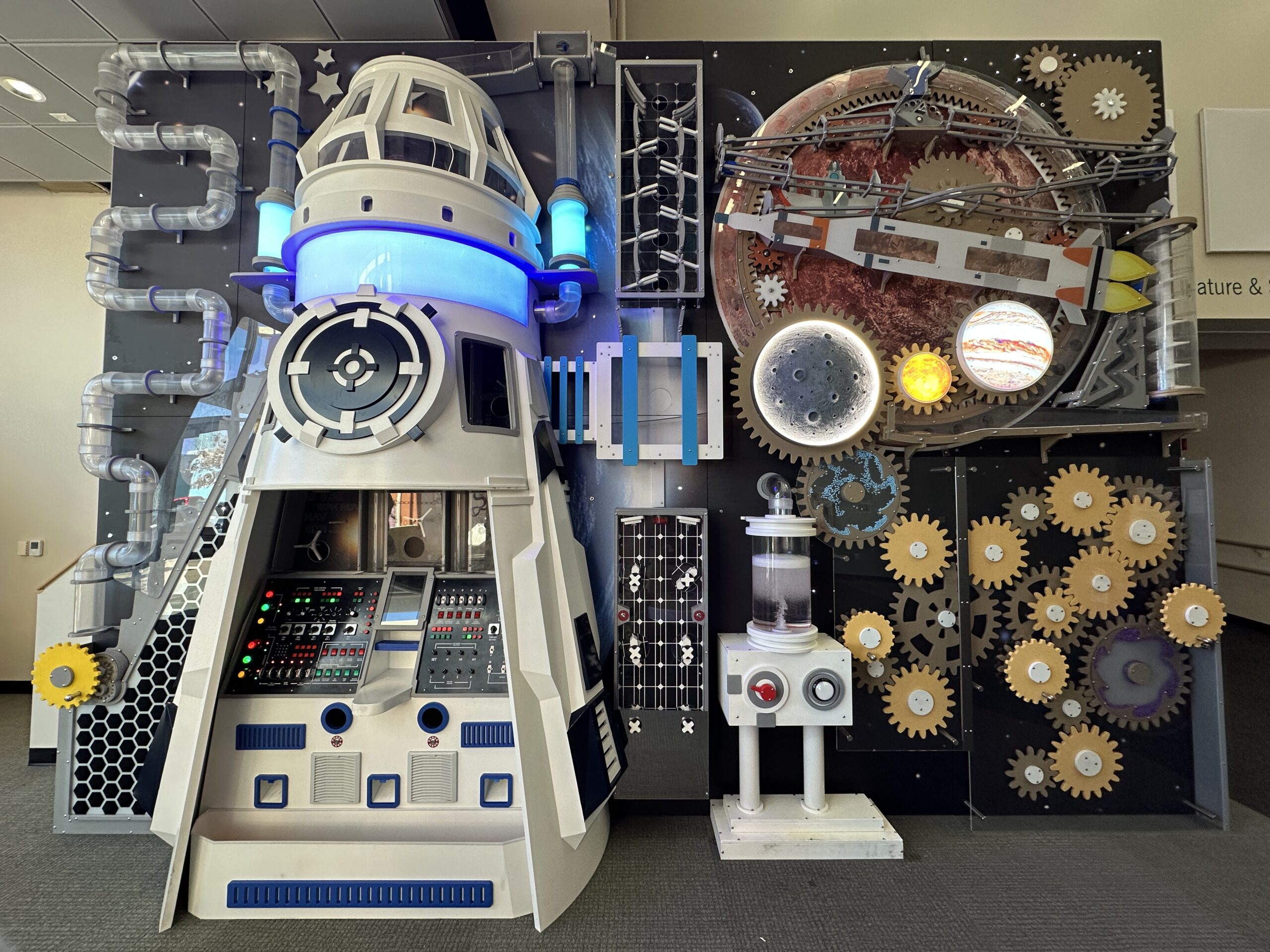 A picture of large, complicated interactive with gears, tubes, wheels, and more in the shape of a spaceship in front of Pluto.