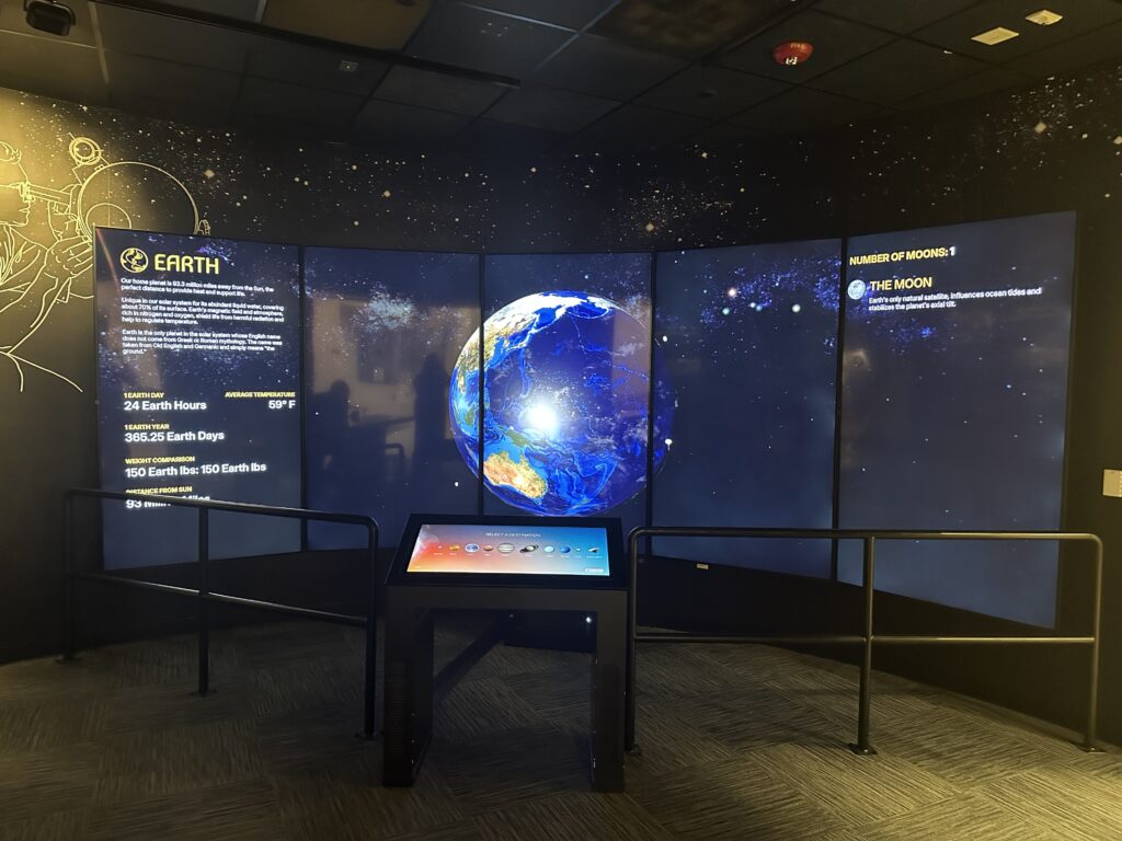 “Journey to Pluto” exhibit at the Museum of Nature & Science. In the photo, it is seen featuring a large multi-panel display of Earth in space, with interactive touchscreen kiosk and educational information about the planet and its moon.