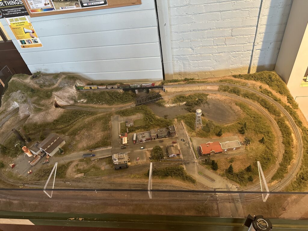 Overhead view of a detailed model railroad scene at the railroad museum, showing miniature trains, tracks, buildings, a water tower, and landscaped hills inside a glass display case.