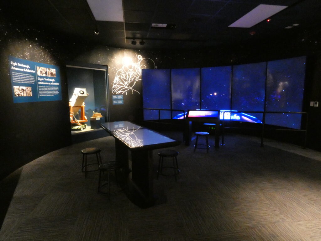 A picture of the Tombaugh room at the Museu of Nature & Science,. It is dark with only the screens on the walls, the Stellar Playground interactive table, and the Grazer Gazer telescope behind glass lluminated.