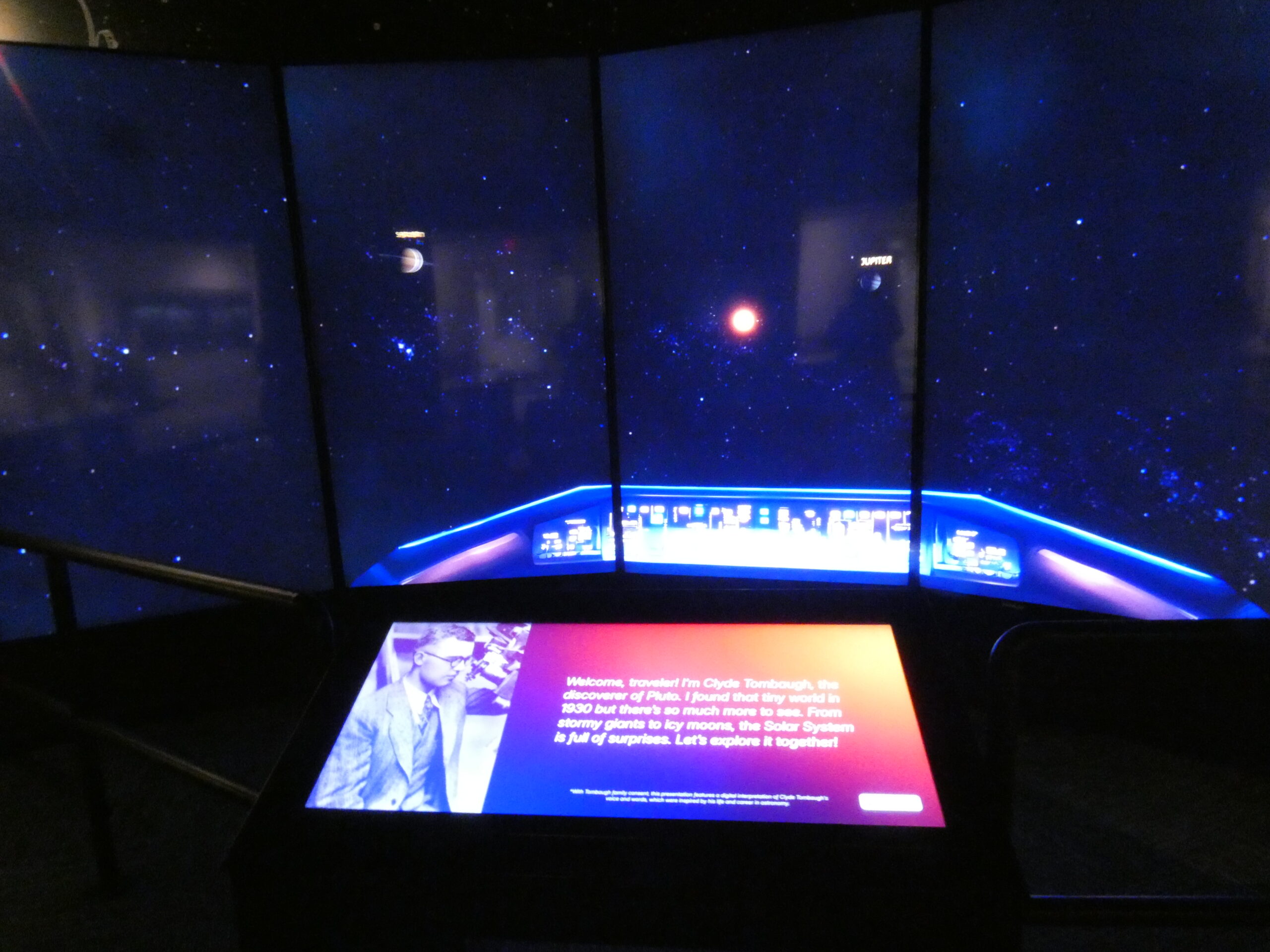 A picture of the Journey to Pluto interactive made of 5 large wall-mounted vertical screens creating a vision of our solar system, with a central console screen with descriptive information on it.