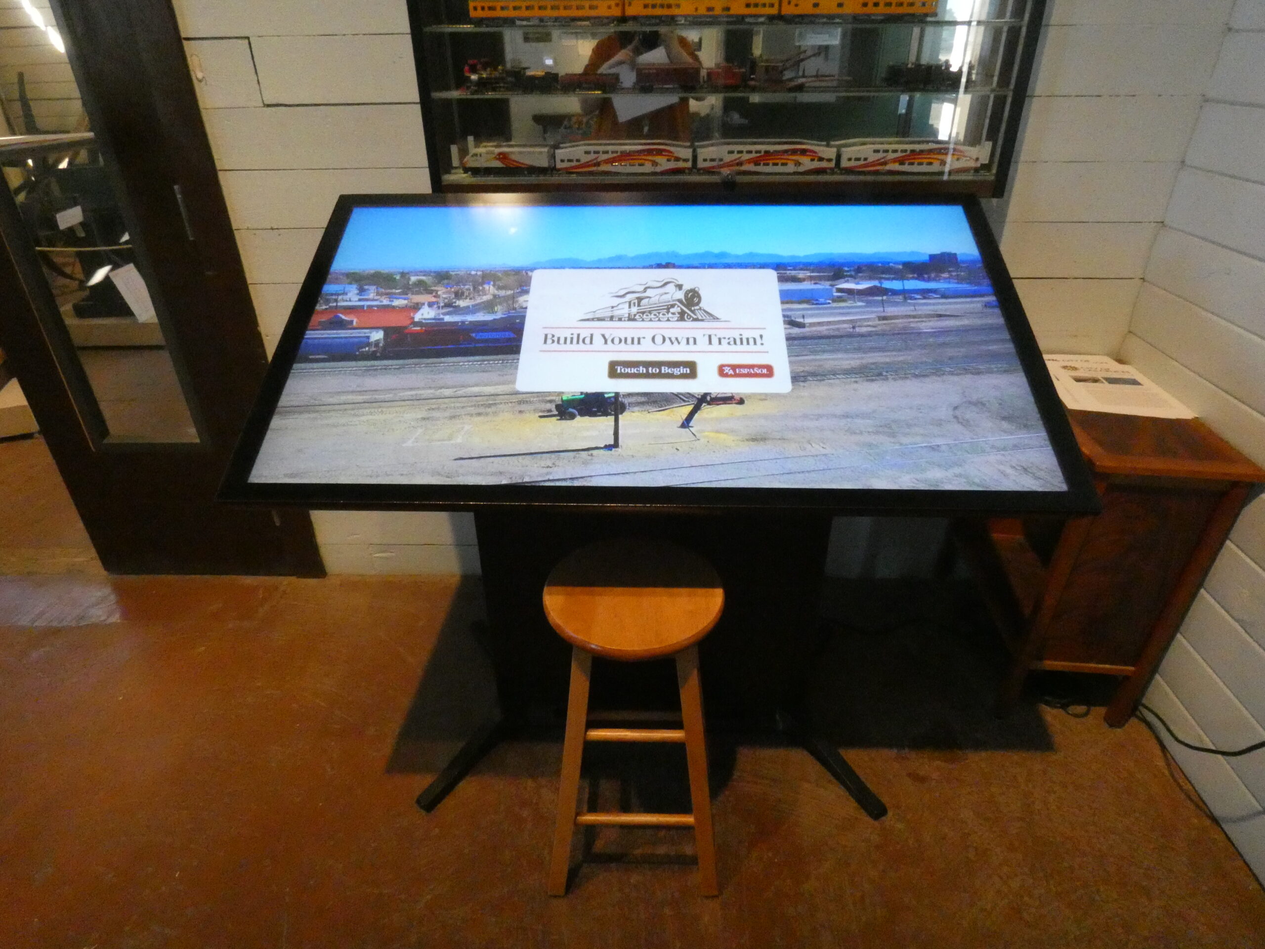 A picture of he "Build Your Own Train!" interactive at the Railroad Museum. It is a large, tiltted screen with a stool in front of it showing title text and trains.