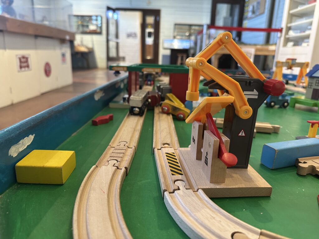 Close-up of a wooden train play table at the railroad museum, featuring toy tracks, small vehicles, and a yellow crane loading area.