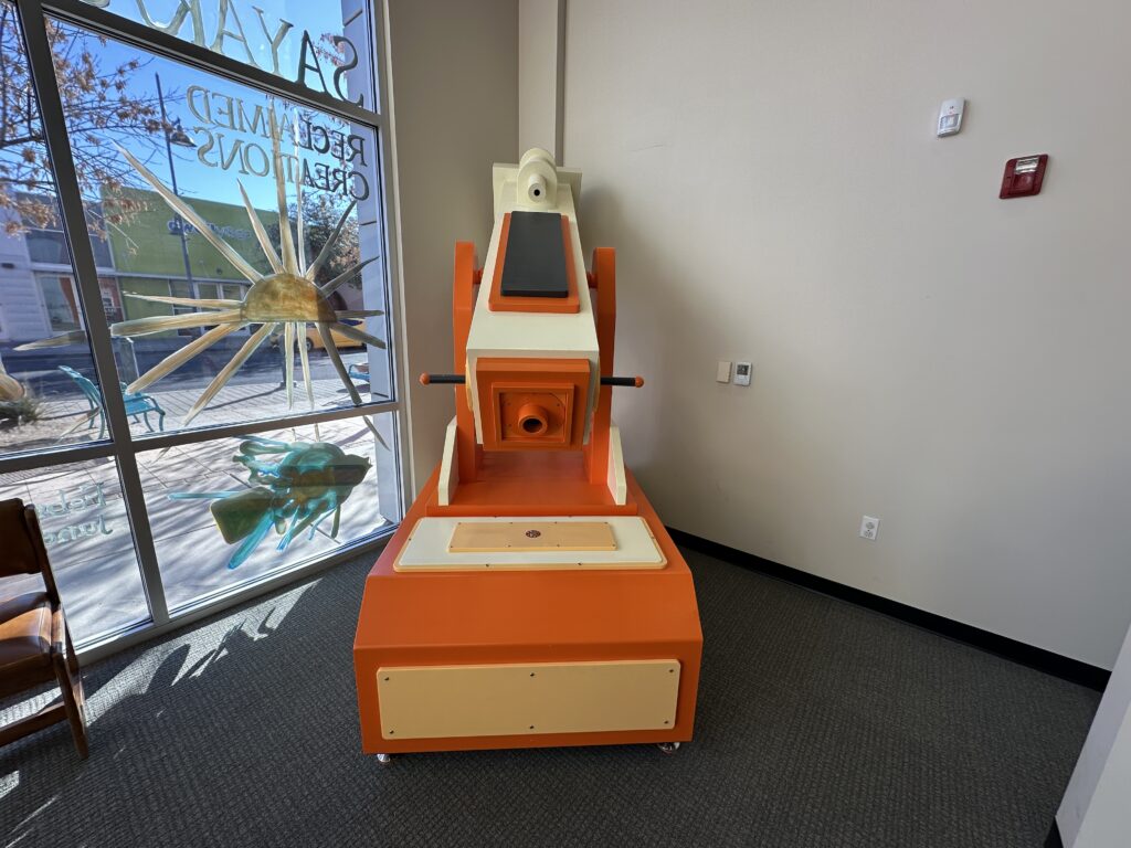 Orange and white interactive telescope exhibit inside the museum, positioned near large windows with painted outdoor-themed designs.