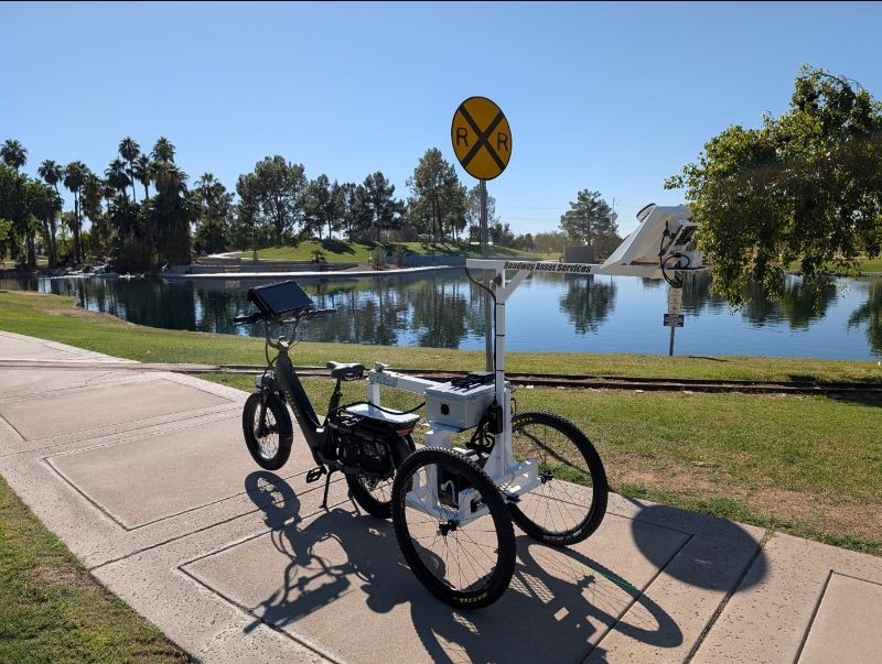 A data collection e-bike parked on the sidewalk of a park with a lake and trees in the background.