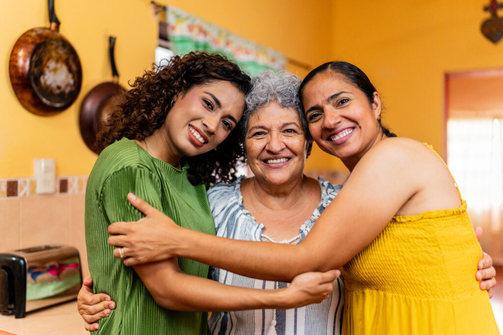 Three women standing in a kitchen hugging each other while looking at the camera.