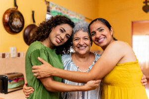 Three women standing in a kitchen hugging each other while looking at the camera.
