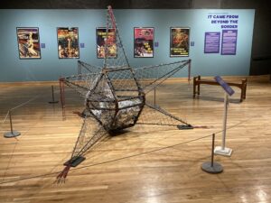 A room at the Branigan Cultural Center displays an exhibit. There is a large metal looking structure on the floor and images in frames on the walls.