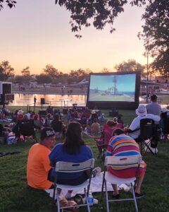 People sitting on the grass at a park watching a movie with a pond in the background.